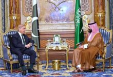 Pakistani Prime Minister Shehbaz Sharif seated on the left in a dark suit, conversing with Saudi Crown Prince Mohammed bin Salman seated on the right in traditional Saudi attire, with the national flags of Pakistan and Saudi Arabia standing between them in an ornate room.