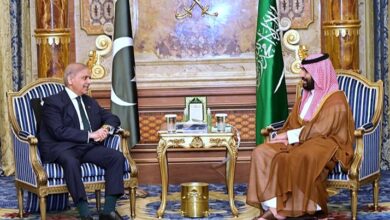 Pakistani Prime Minister Shehbaz Sharif seated on the left in a dark suit, conversing with Saudi Crown Prince Mohammed bin Salman seated on the right in traditional Saudi attire, with the national flags of Pakistan and Saudi Arabia standing between them in an ornate room.