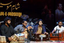 Musicians performing live on stage at a Sham-e-Moseeqi musical event at PNCA, with a harmonium, tabla, and traditional string instruments, under a backdrop featuring Urdu calligraphy.