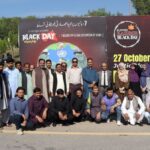 NBF Rallies – Group of National Book Foundation (NBF) male and female officials and staff standing and crouching together outdoors in front of a large red and black Kashmir Black Day awareness backdrop with Urdu and English text on 27 October.