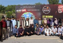 NBF Rallies – Group of National Book Foundation (NBF) male and female officials and staff standing and crouching together outdoors in front of a large red and black Kashmir Black Day awareness backdrop with Urdu and English text on 27 October.