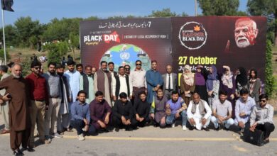 NBF Rallies – Group of National Book Foundation (NBF) male and female officials and staff standing and crouching together outdoors in front of a large red and black Kashmir Black Day awareness backdrop with Urdu and English text on 27 October.