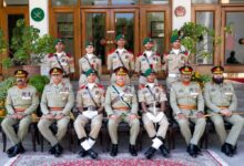 A formal group photograph posing outdoors on steps in front of a wooden building entry. Sitting in the front row are senior military officers in light olive-green parade uniforms decorated with medals, shoulder boards, and sashes, alternating with three award-winning military cadets in light-khaki drill uniforms wearing white gloves and white spats over black boots. In the centre sits the Chief Guest officer, flanked by cadets holding ceremonial swords and canes. Standing in the back row are five additional cadets in matching light-khaki uniforms with green berets, red epaulettes, and chest ribbons. A patterned red carpet is laid out on the stone pavement in front of the seating arrangement.