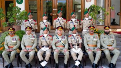 A formal group photograph posing outdoors on steps in front of a wooden building entry. Sitting in the front row are senior military officers in light olive-green parade uniforms decorated with medals, shoulder boards, and sashes, alternating with three award-winning military cadets in light-khaki drill uniforms wearing white gloves and white spats over black boots. In the centre sits the Chief Guest officer, flanked by cadets holding ceremonial swords and canes. Standing in the back row are five additional cadets in matching light-khaki uniforms with green berets, red epaulettes, and chest ribbons. A patterned red carpet is laid out on the stone pavement in front of the seating arrangement.