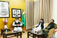 The KP Governor visits Al-Markaz-ul-Islami Peshawar. A wide interior photograph depicting a high-level political meeting inside a paneled office setting. On the left, Governor Khyber Pakhtunkhwa Faisal Karim Kundi, wearing a white shalwar kameez and dark waistcoat, gestures while speaking from a wooden armchair. Seated at the head of the room is Professor Muhammad Ibrahim Khan, wearing a traditional turban, vest, and white attire. On the right side, three senior Jamaat-e-Islami leaders sit on a sofa listening attentively. The room features wood-panelled walls adorned with framed calligraphy, a scale of justice emblem, and a Pakistani flag. Glass coffee tables set with water bottles and floral arrangements sit in front of the participants.