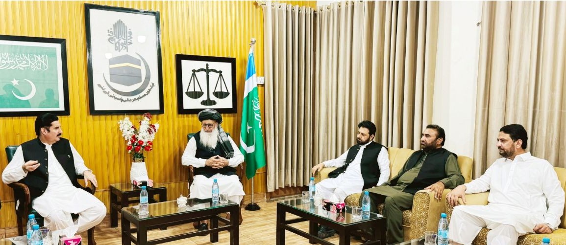 The KP Governor visits Al-Markaz-ul-Islami Peshawar. A wide interior photograph depicting a high-level political meeting inside a paneled office setting. On the left, Governor Khyber Pakhtunkhwa Faisal Karim Kundi, wearing a white shalwar kameez and dark waistcoat, gestures while speaking from a wooden armchair. Seated at the head of the room is Professor Muhammad Ibrahim Khan, wearing a traditional turban, vest, and white attire. On the right side, three senior Jamaat-e-Islami leaders sit on a sofa listening attentively. The room features wood-panelled walls adorned with framed calligraphy, a scale of justice emblem, and a Pakistani flag. Glass coffee tables set with water bottles and floral arrangements sit in front of the participants.