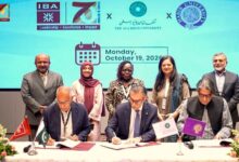AI in Pedagogy: A group photograph taken during a formal signing ceremony on stage. Three male university leaders sit side by side at a draped table, each signing official documents placed before small desk flags representing their respective institutions and Pakistan. Standing directly behind them are five academics and officials, three women and two men, smiling as witnesses. Decorative floral arrangements frame the left and right edges of the desk. A large digital backdrop displays the institutional logos of IBA Karachi, Aga Khan University, and Habib University alongside a calendar graphic indicating. Monday, October 19, 2026.
