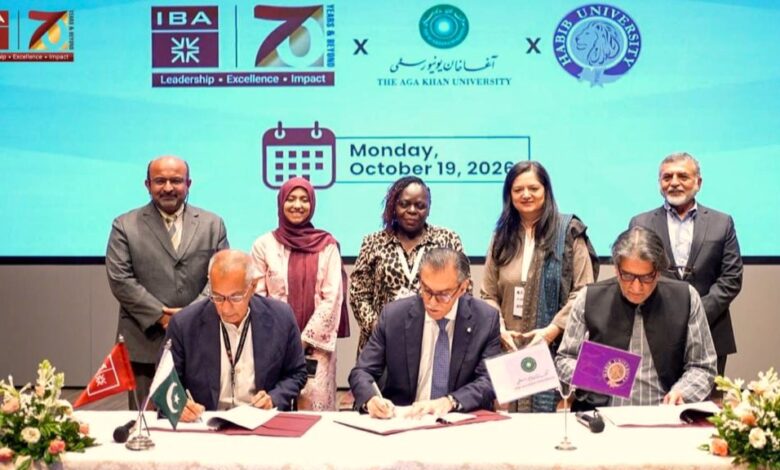 AI in Pedagogy: A group photograph taken during a formal signing ceremony on stage. Three male university leaders sit side by side at a draped table, each signing official documents placed before small desk flags representing their respective institutions and Pakistan. Standing directly behind them are five academics and officials, three women and two men, smiling as witnesses. Decorative floral arrangements frame the left and right edges of the desk. A large digital backdrop displays the institutional logos of IBA Karachi, Aga Khan University, and Habib University alongside a calendar graphic indicating. Monday, October 19, 2026.
