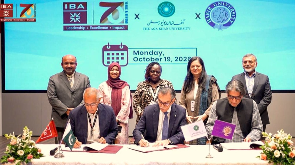 AI in Pedagogy: A group photograph taken during a formal signing ceremony on stage. Three male university leaders sit side by side at a draped table, each signing official documents placed before small desk flags representing their respective institutions and Pakistan. Standing directly behind them are five academics and officials, three women and two men, smiling as witnesses. Decorative floral arrangements frame the left and right edges of the desk. A large digital backdrop displays the institutional logos of IBA Karachi, Aga Khan University, and Habib University alongside a calendar graphic indicating. Monday, October 19, 2026.