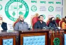 Hina Javed Murder Case: Five individuals are seated behind a large, dark wooden press table lined with multiple news microphones and black cables. In the centre, a man with glasses and a black shirt speaks directly into the microphones. To his right sits a bald man with a beard wearing a grey shirt, and on the far left, an older man wearing a white face mask and a dark waistcoat over a white shalwar kameez. To the speaker’s left sits a woman wearing a black face mask and headscarf, alongside another woman in a patterned yellow and brown outfit. Behind them, high-backed red upholstered chairs are set against a white backdrop featuring green and black "National Press Club Islamabad" logos and branding.