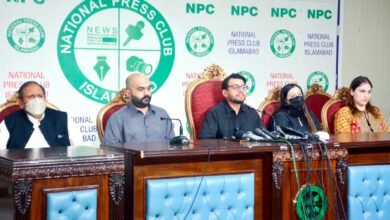 Hina Javed Murder Case: Five individuals are seated behind a large, dark wooden press table lined with multiple news microphones and black cables. In the centre, a man with glasses and a black shirt speaks directly into the microphones. To his right sits a bald man with a beard wearing a grey shirt, and on the far left, an older man wearing a white face mask and a dark waistcoat over a white shalwar kameez. To the speaker’s left sits a woman wearing a black face mask and headscarf, alongside another woman in a patterned yellow and brown outfit. Behind them, high-backed red upholstered chairs are set against a white backdrop featuring green and black "National Press Club Islamabad" logos and branding.