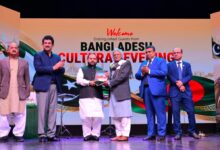 Pakistan and Bangladesh: Five men in formal attire stand on a brightly lit stage in front of a colorful backdrop that reads "Welcome Distinguished Guests from Bangladesh Cultural Evening" alongside Pakistani and Bangladeshi flags. On the left, Aurangzeb Khan Khichi applauds while wearing a navy blazer and trousers, alongside a man in a brown jacket and shalwar kameez. In the center, Director General Muhammad Ayoub Jamali (in a waistcoat and white shalwar kameez) hands a commemorative shield to an elderly Bangladeshi delegate in a grey coat and scarf. To the right, Secretary Asad Ur Rahman Gillani (in a bright blue suit) and Bangladesh High Commissioner Md. Iqbal Hussain Khan (in a dark blue suit) stand applauding. Stage equipment, microphone stands, and the colorful "Welcome" graphics are visible on the stage and backdrop.