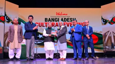Pakistan and Bangladesh: Five men in formal attire stand on a brightly lit stage in front of a colorful backdrop that reads "Welcome Distinguished Guests from Bangladesh Cultural Evening" alongside Pakistani and Bangladeshi flags. On the left, Aurangzeb Khan Khichi applauds while wearing a navy blazer and trousers, alongside a man in a brown jacket and shalwar kameez. In the center, Director General Muhammad Ayoub Jamali (in a waistcoat and white shalwar kameez) hands a commemorative shield to an elderly Bangladeshi delegate in a grey coat and scarf. To the right, Secretary Asad Ur Rahman Gillani (in a bright blue suit) and Bangladesh High Commissioner Md. Iqbal Hussain Khan (in a dark blue suit) stand applauding. Stage equipment, microphone stands, and the colorful "Welcome" graphics are visible on the stage and backdrop.