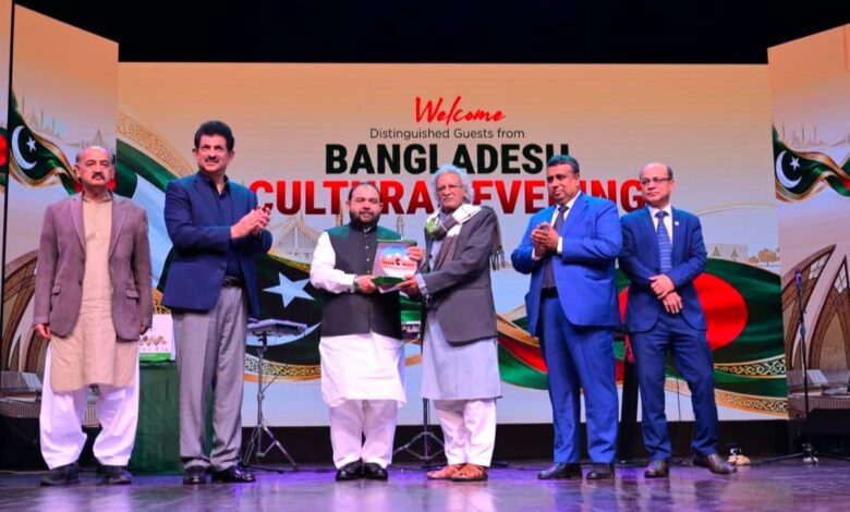 Pakistan and Bangladesh: Five men in formal attire stand on a brightly lit stage in front of a colorful backdrop that reads "Welcome Distinguished Guests from Bangladesh Cultural Evening" alongside Pakistani and Bangladeshi flags. On the left, Aurangzeb Khan Khichi applauds while wearing a navy blazer and trousers, alongside a man in a brown jacket and shalwar kameez. In the center, Director General Muhammad Ayoub Jamali (in a waistcoat and white shalwar kameez) hands a commemorative shield to an elderly Bangladeshi delegate in a grey coat and scarf. To the right, Secretary Asad Ur Rahman Gillani (in a bright blue suit) and Bangladesh High Commissioner Md. Iqbal Hussain Khan (in a dark blue suit) stand applauding. Stage equipment, microphone stands, and the colorful "Welcome" graphics are visible on the stage and backdrop.