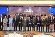 SVI Convenes Landmark: A large group of over thirty international delegates, scholars, and military officials standing together in rows on a carpeted stage inside a formal conference hall. The men are dressed predominantly in formal dark business suits and ties, while several female attendees wear business attire or vibrant traditional Pakistani wear. Behind the group is a wide backdrop displaying the Strategic Vision Institute logo and text that reads "ISLAMABAD NON-PROLIFERATION CONFERENCE INC-2026". Large digital displays flanking the main backdrop show the conference branding with "DAY 01" prominently featured on the left panel. In the foreground, round tables set with water bottles and floral arrangements are visible.