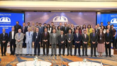 SVI Convenes Landmark: A large group of over thirty international delegates, scholars, and military officials standing together in rows on a carpeted stage inside a formal conference hall. The men are dressed predominantly in formal dark business suits and ties, while several female attendees wear business attire or vibrant traditional Pakistani wear. Behind the group is a wide backdrop displaying the Strategic Vision Institute logo and text that reads "ISLAMABAD NON-PROLIFERATION CONFERENCE INC-2026". Large digital displays flanking the main backdrop show the conference branding with "DAY 01" prominently featured on the left panel. In the foreground, round tables set with water bottles and floral arrangements are visible.