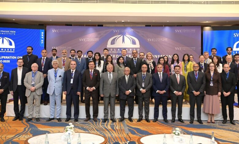 SVI Convenes Landmark: A large group of over thirty international delegates, scholars, and military officials standing together in rows on a carpeted stage inside a formal conference hall. The men are dressed predominantly in formal dark business suits and ties, while several female attendees wear business attire or vibrant traditional Pakistani wear. Behind the group is a wide backdrop displaying the Strategic Vision Institute logo and text that reads "ISLAMABAD NON-PROLIFERATION CONFERENCE INC-2026". Large digital displays flanking the main backdrop show the conference branding with "DAY 01" prominently featured on the left panel. In the foreground, round tables set with water bottles and floral arrangements are visible.